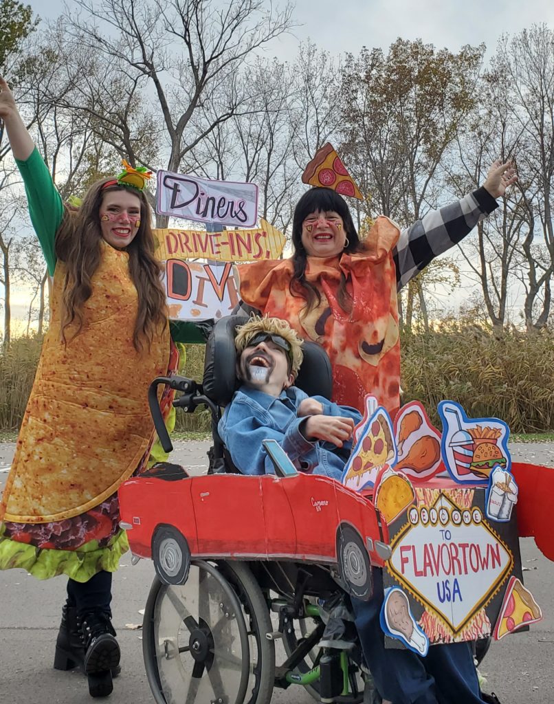 women in taco and pizza slice costumes stand next to man dressed like Guy Fieri in wheelchair decorated like red car