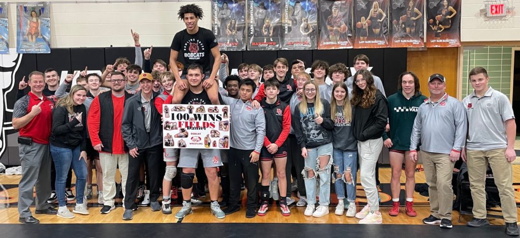 Wrestler sits on shoulders of man surrounded by teammates with 100 wins sign