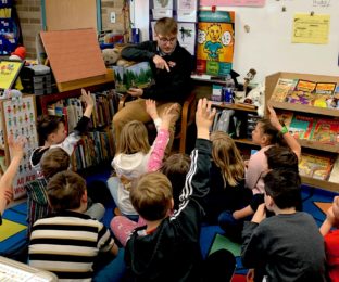 Man holds book sitting in chair; children face him with hands raised to answer questions