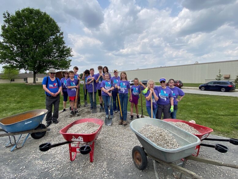 15 people stand with shovels in front of three wheelbarrows filled with landscape stones