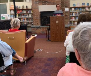 Man at podium talks to women in audience at public library.