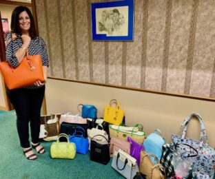 Woman stands holding large orange purse, variety of purses sit on floor to the right.
