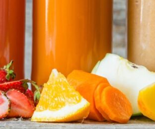 Red, pink, orange, yellow, green smoothies in glasses; blackberries, blueberries, raspberries, strawberries, oranges, carrots, peaches, bananas and spinach in front of glasses