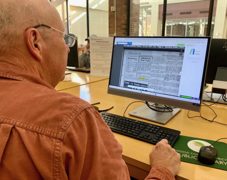 Computer screen is seen over the shoulder of man looking at a historical newspaper website.