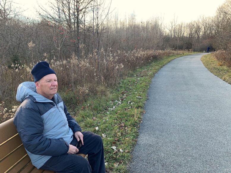 Man sits on park bench in late fall; paved trail on the right.