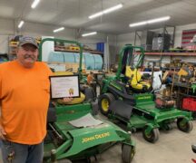 Jim Blackford holds plaque in maintenance shop with John Deere tractors, tools and other equipment.