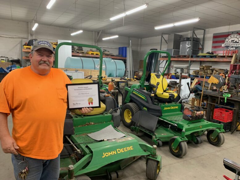 Jim Blackford holds plaque in maintenance shop with John Deere tractors, tools and other equipment.