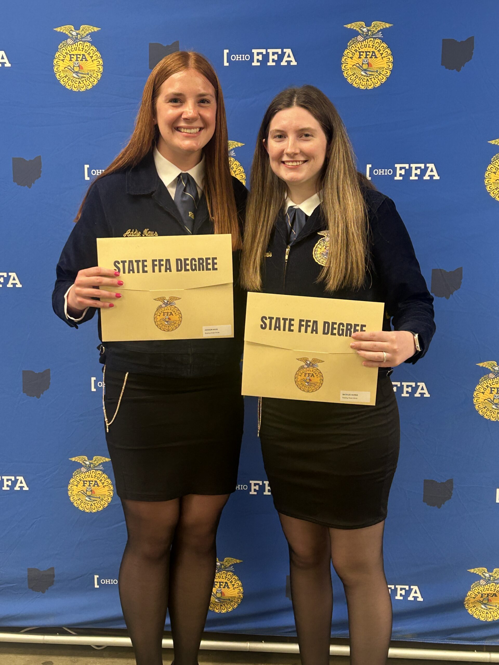 Two high school girls, State Degree Recipients Addie Naus and Natalee Harris, stand in front of a blue backdrop, holding their degree certificates.