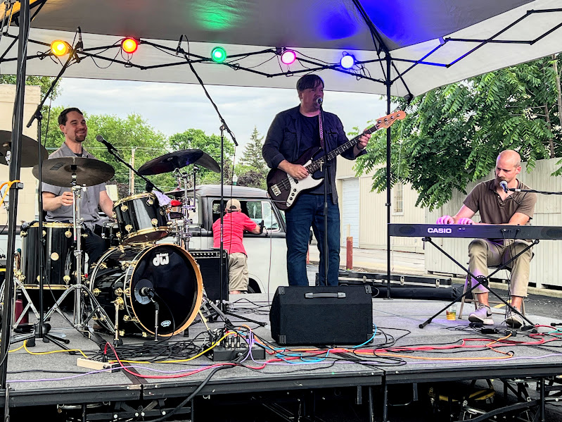 three people perform on stage: one is sitting and drumming, another is standing and playing guitar, while the third is sitting and playing keyboard