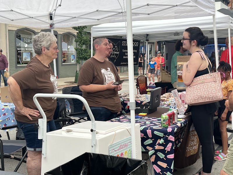 two women in brown shirts stand behind a table while one speaks to a girl across the table from them