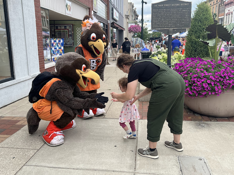 The photo shows two mascots, falcons, greeting a young toddler and her mother. One of the mascots is squatting to match the height of the child.