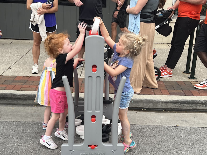 two girls stand on either side of a large connect four game - each girl is trying to put a piece in the game