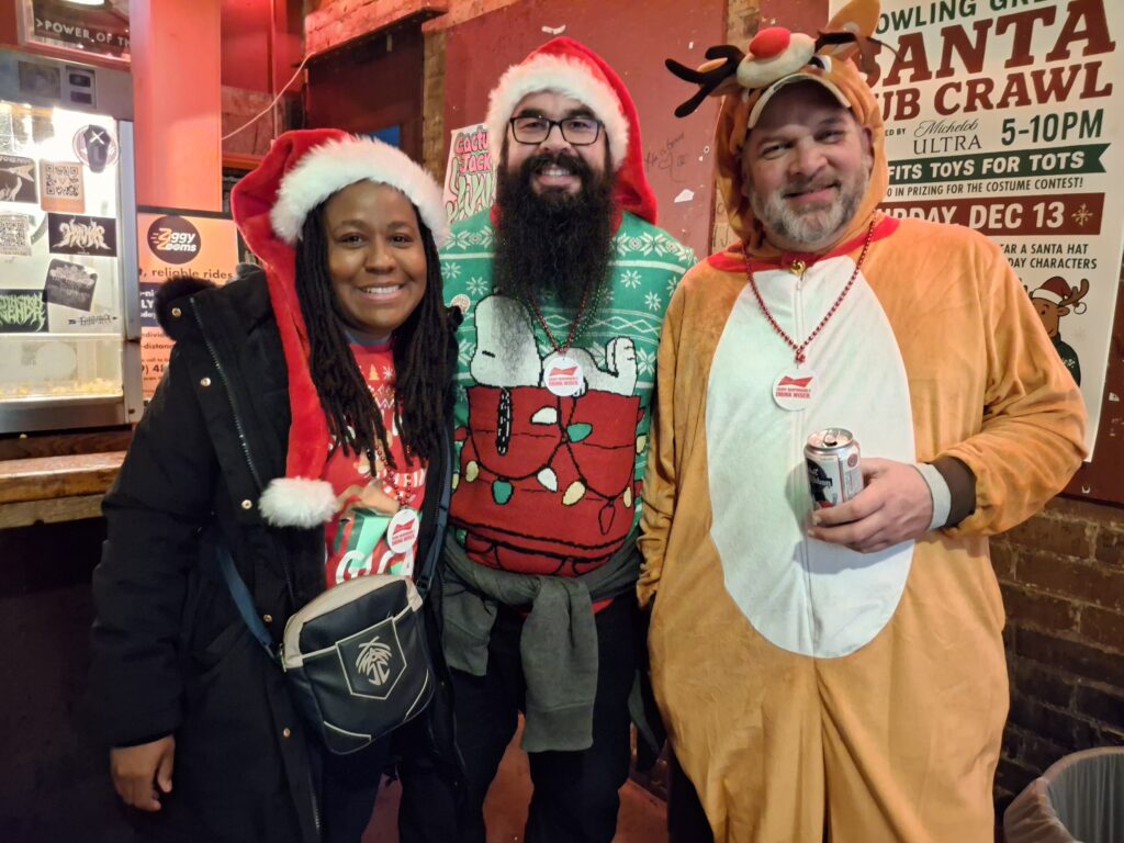 Ashley Wynn, Brian Urias and Sean Brennan wearing Christmas sweaters and a reindeer costume.