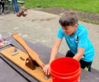A young boy in a turquois athletic shirt is outside and uses a table-top catapult to launch a stuffed animal into a bucket.