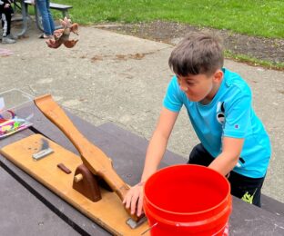 A young boy in a turquois athletic shirt is outside and uses a table-top catapult to launch a stuffed animal into a bucket.