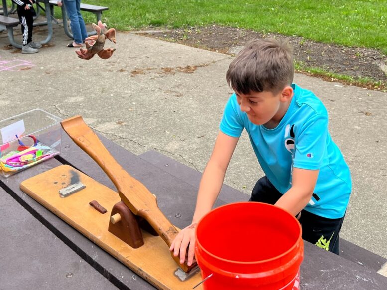 A young boy in a turquois athletic shirt is outside and uses a table-top catapult to launch a stuffed animal into a bucket.