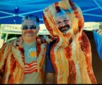 Two men with outfits featuring bacon smile under a festival tent.