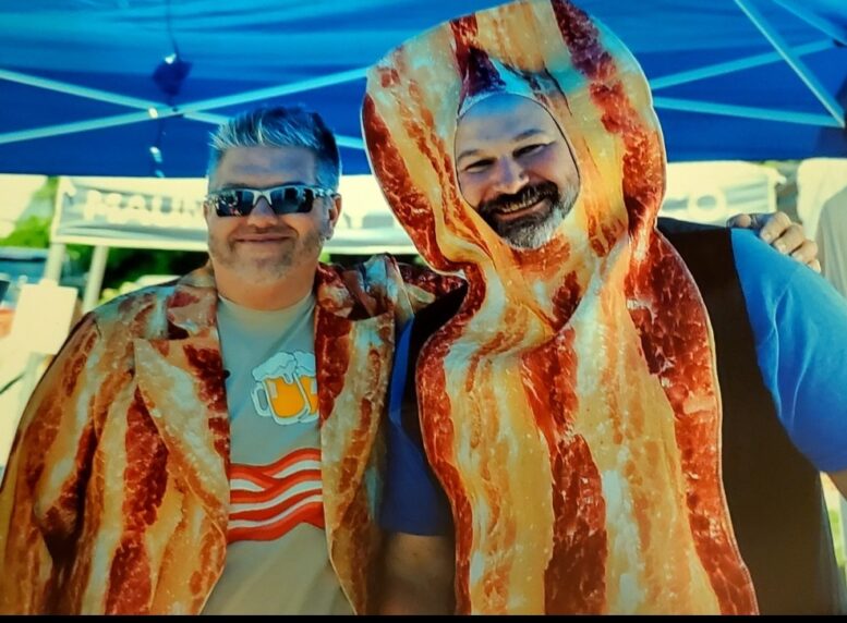 Two men with outfits featuring bacon smile under a festival tent.