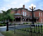 Picture featuring a large historic brick building, which is a museum, a blue sky, and a wrought iron fence.