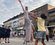 a performer walks down the street on stilts, blowing bubbles while a young girl jumps to grab the bubbles