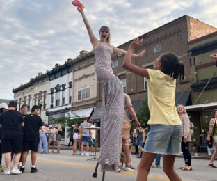 a performer walks down the street on stilts, blowing bubbles while a young girl jumps to grab the bubbles
