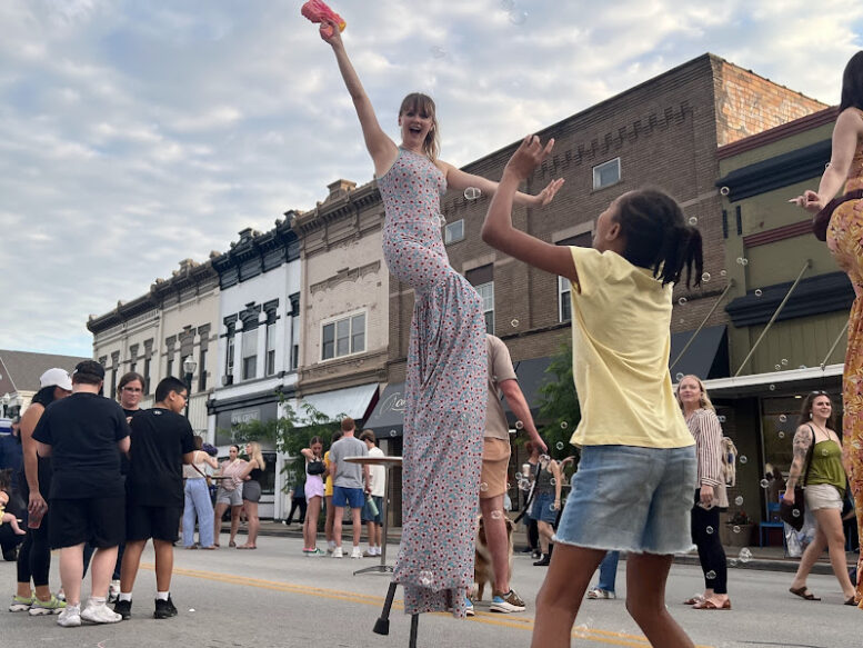 a performer walks down the street on stilts, blowing bubbles while a young girl jumps to grab the bubbles
