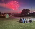 Group of teenagers sits in the grass and watches as a red firework explodes in the sky