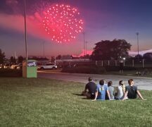 Group of teenagers sits in the grass and watches as a red firework explodes in the sky