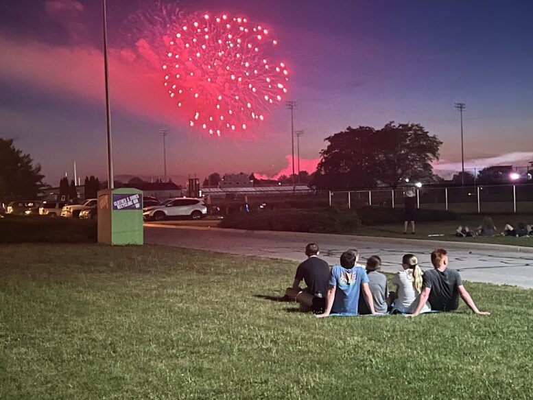 Group of teenagers sits in the grass and watches as a red firework explodes in the sky