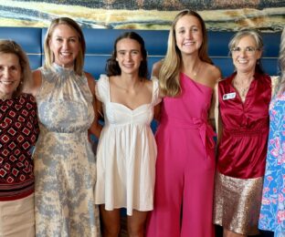 six women wearing dressier clothes smile at the camera with their arms around each other
