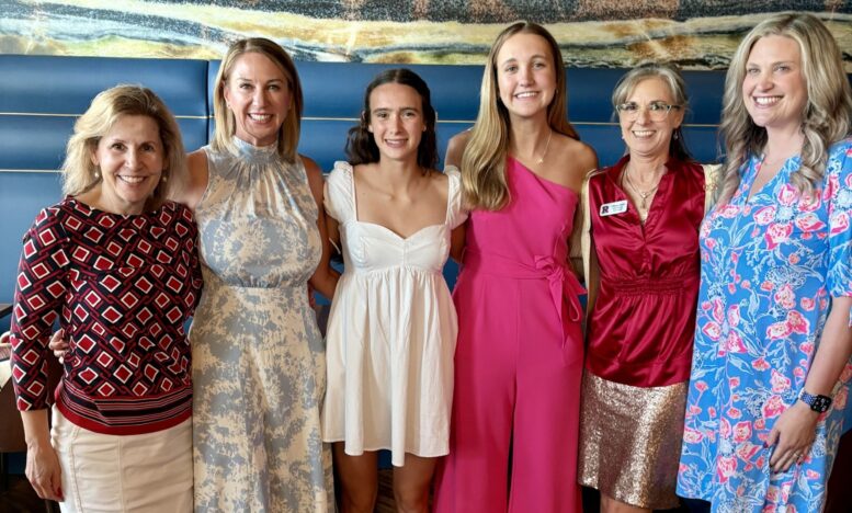 six women wearing dressier clothes smile at the camera with their arms around each other
