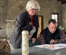 Woman stands at table; man sits, look at map on the table.