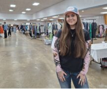 Young woman with long dark hair in a ball cap stands in front of rows of used clothing.