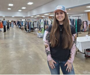 Young woman with long dark hair in a ball cap stands in front of rows of used clothing.