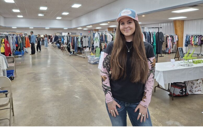 Young woman with long dark hair in a ball cap stands in front of rows of used clothing.