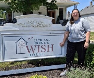 Woman in black pants and gray shirt in front of a sign reading Jack & Irene's WISH House