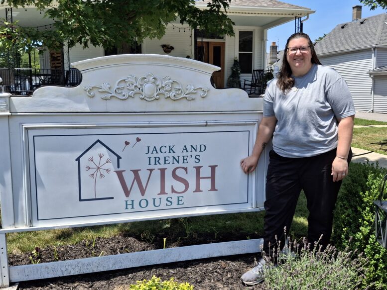 Woman in black pants and gray shirt in front of a sign reading Jack & Irene's WISH House